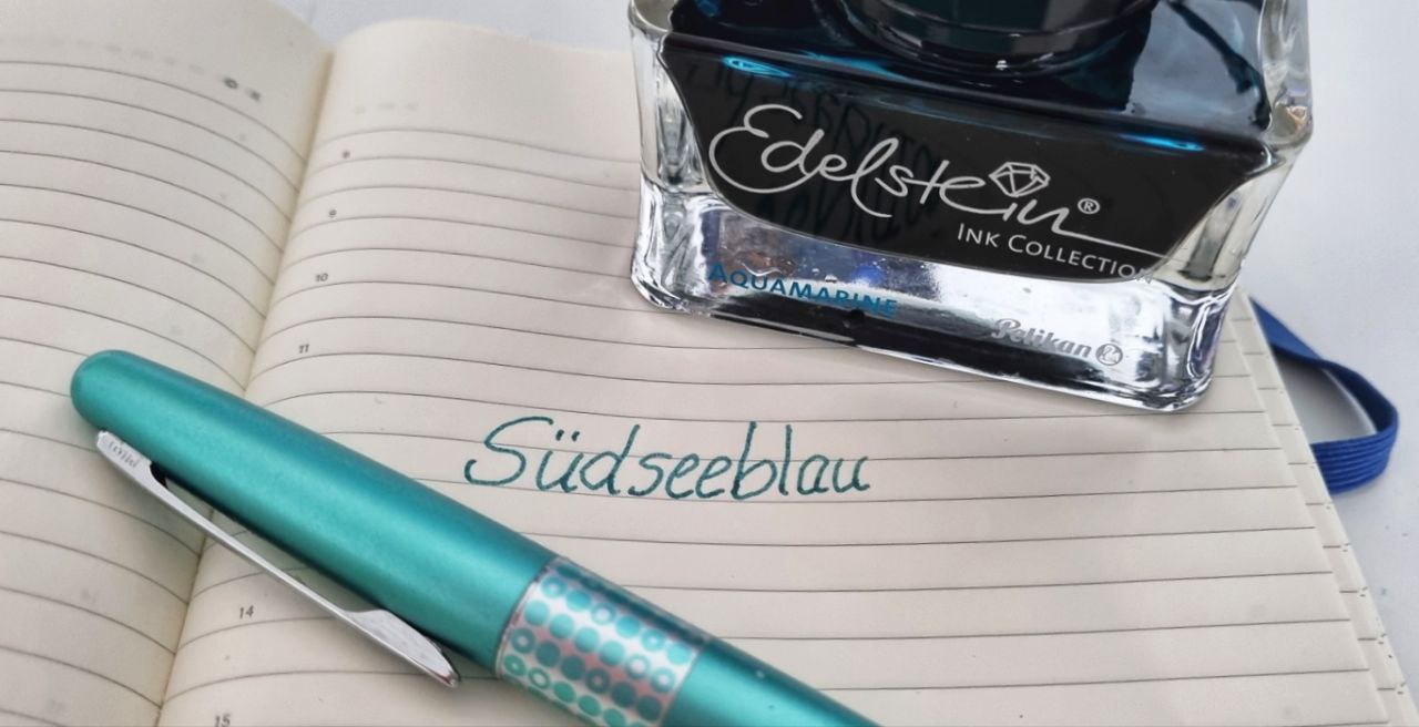 das Wort "Südseeblau" in türkiser Tinte geschrieben, davor ein türkiser Füller, dahinter ein Tintenglas